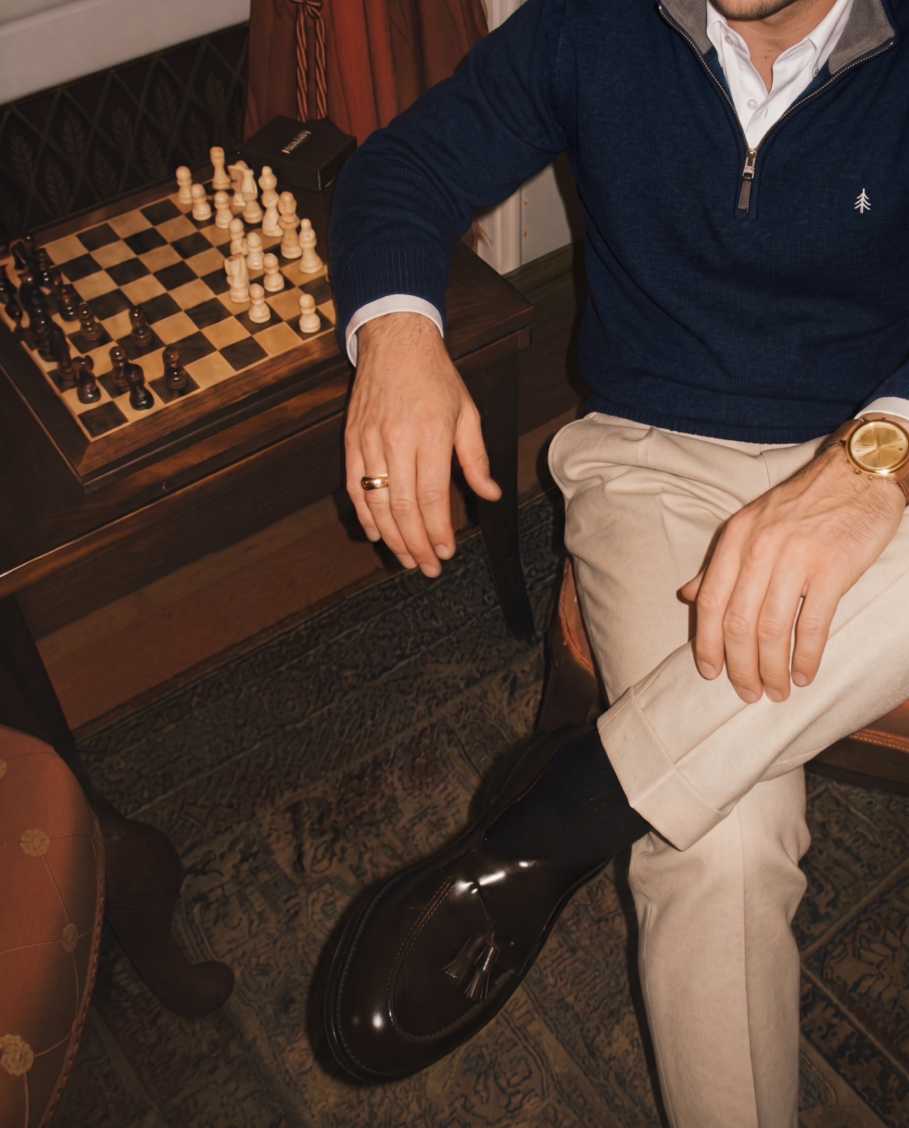 A man seated in a warm panelled room in a navy Cedar Rowe quarter-zip, a chessboard and tassel loafers beside him.