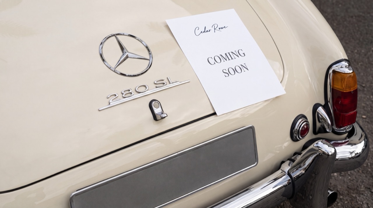 The boot of a vintage cream Mercedes 280SL with a handwritten Cedar Rowe card reading Coming Soon.