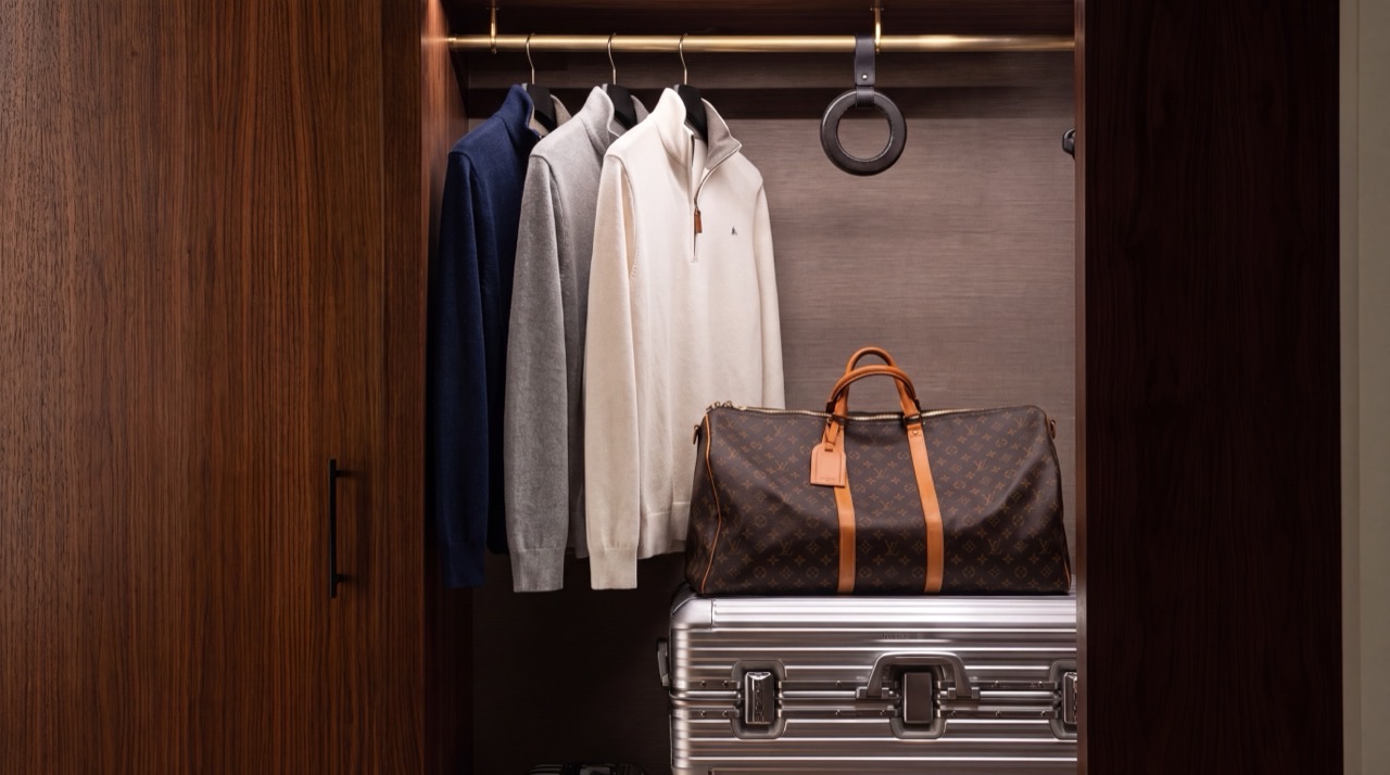 A walnut wardrobe in low light, Cedar Rowe knits in navy, grey and cream on a brass rail beside leather and aluminium luggage.